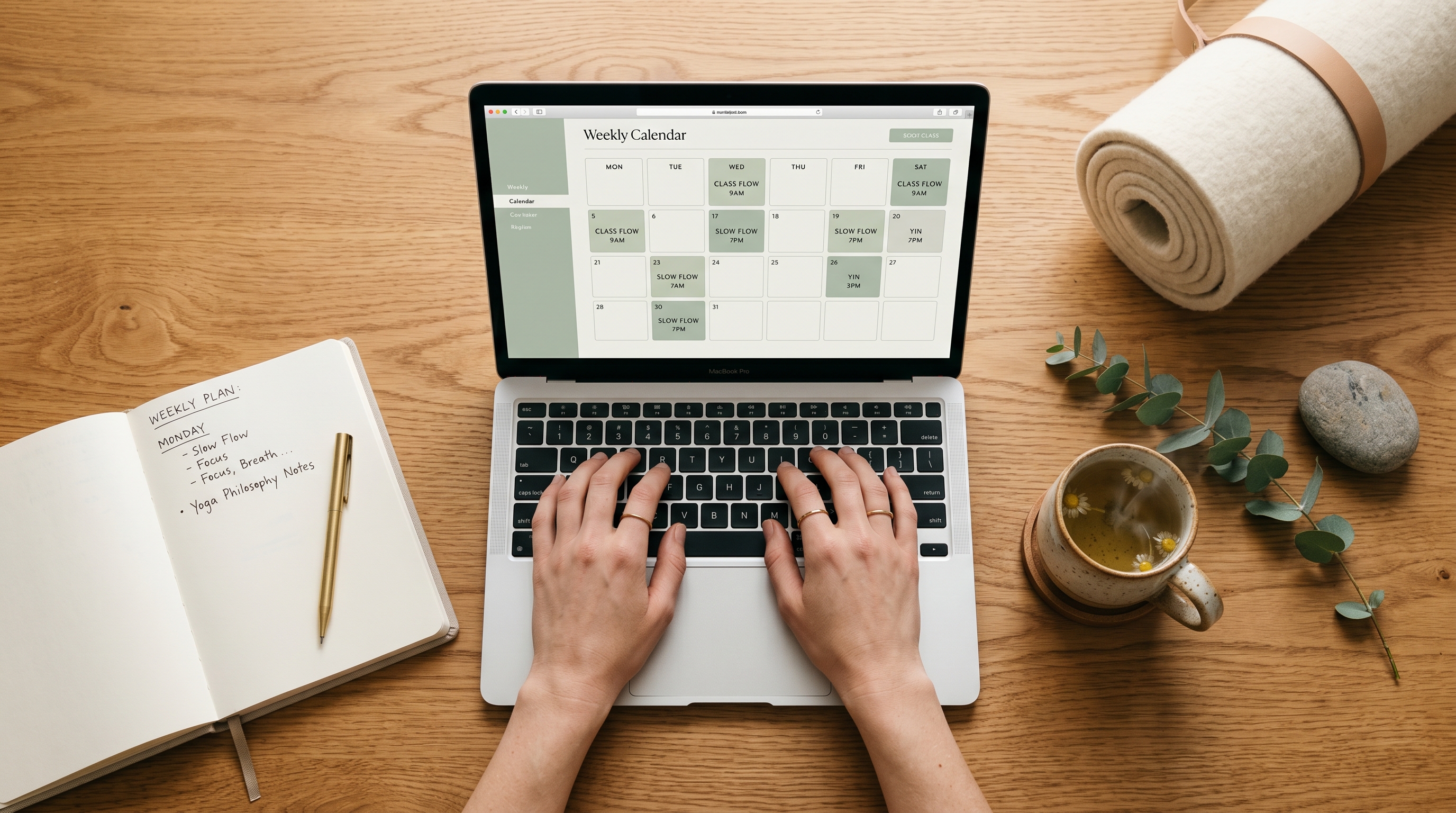 A wellness teacher's workspace — laptop with class calendar, yoga mat, chamomile tea, eucalyptus